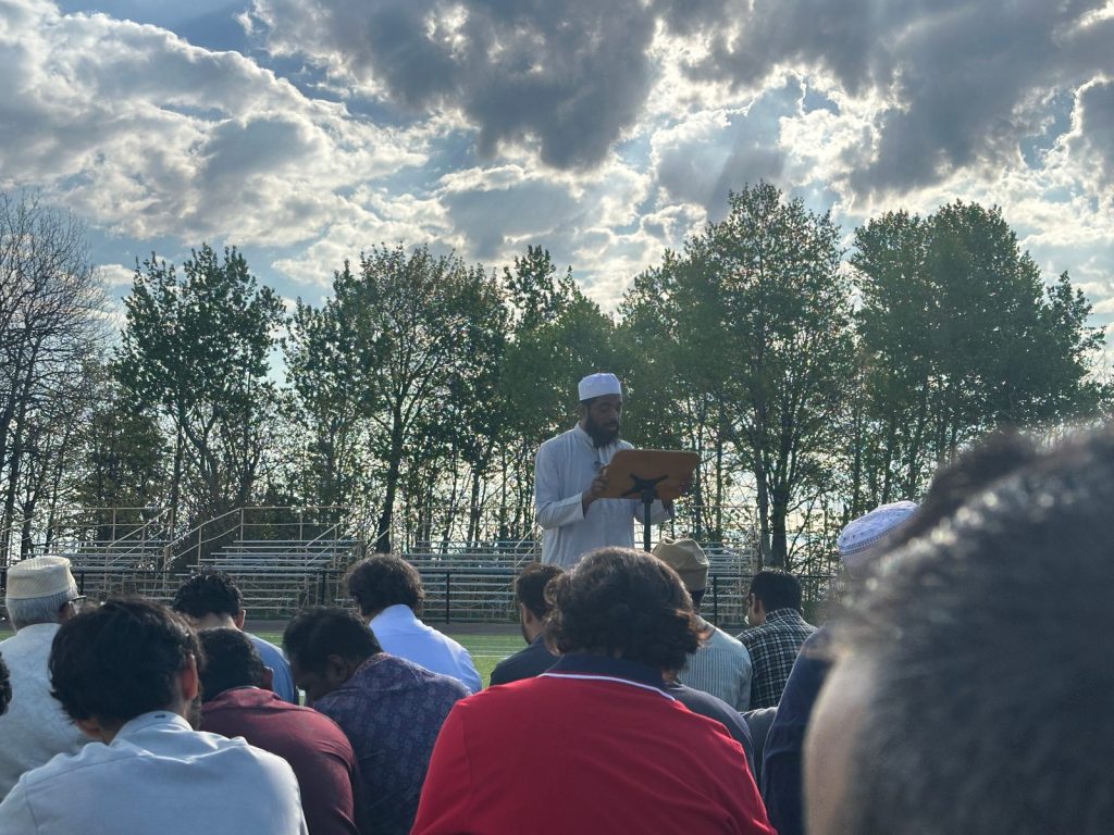 A man in a white cap reads from a lectern as the seated congregation listens.