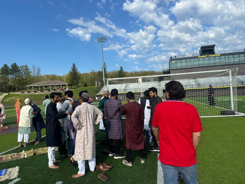 Men in Eid clothes gather and greet each other on the field in front of Kearly Stadium.