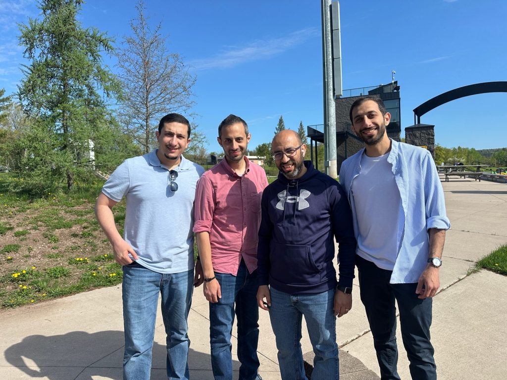 Four smiling men pose together on a sunny path outside the stadium.