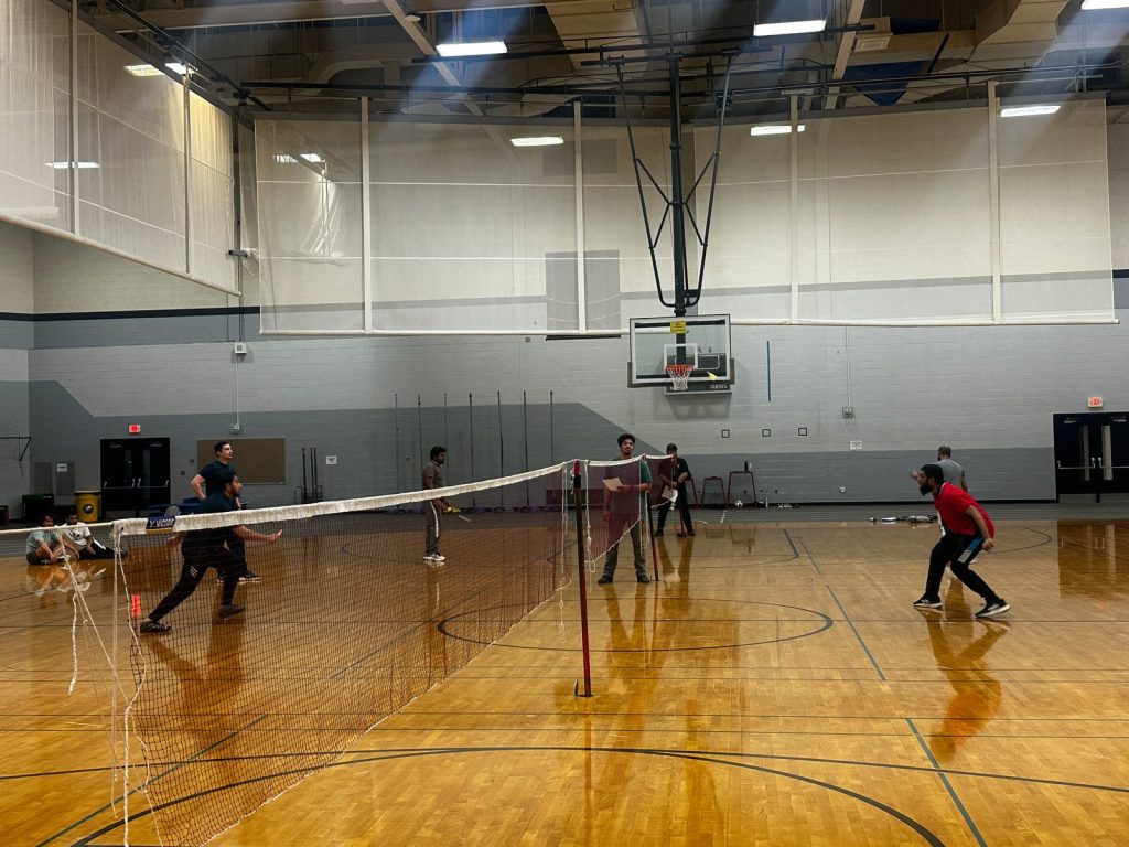Players rally across a badminton net while others watch from beside the court.