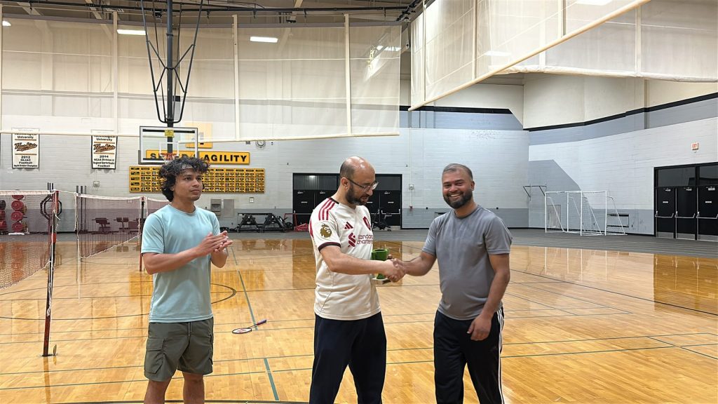 Two men shake hands over the trophy as a third player applauds.