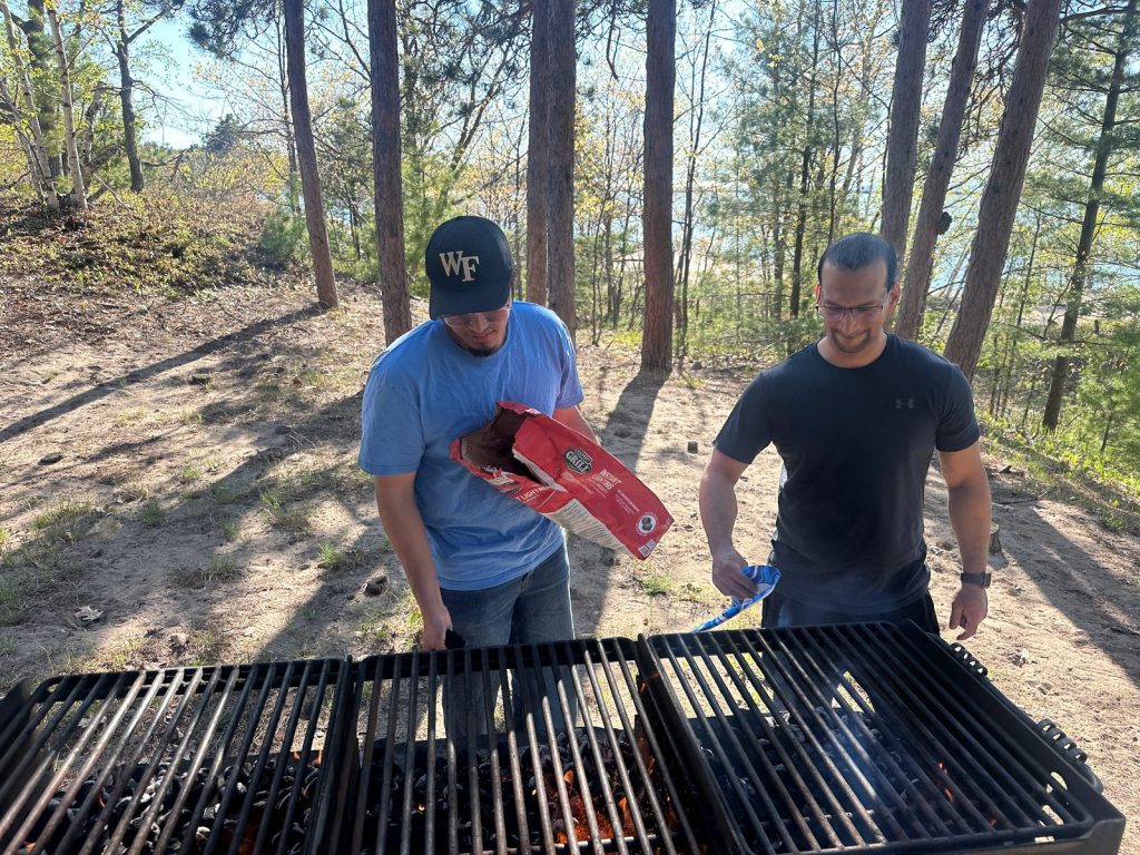 Two men pour charcoal into a row of park grills under the pines.