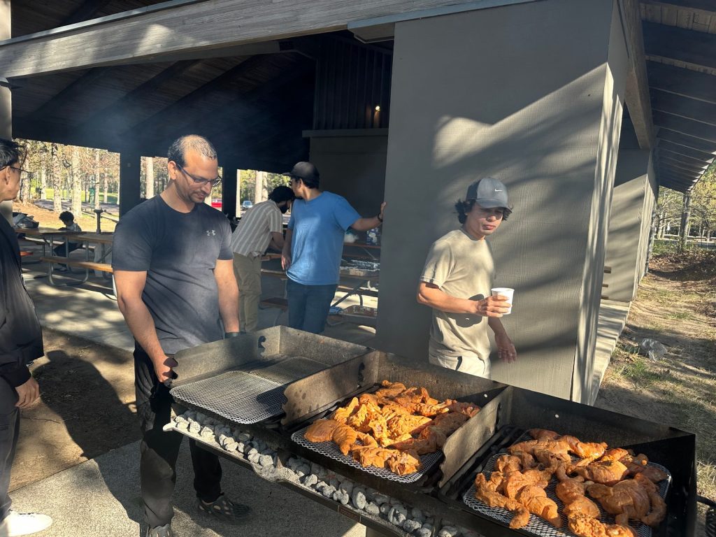 Raw marinated chicken sits on a grill beside the picnic shelter as men stand nearby.
