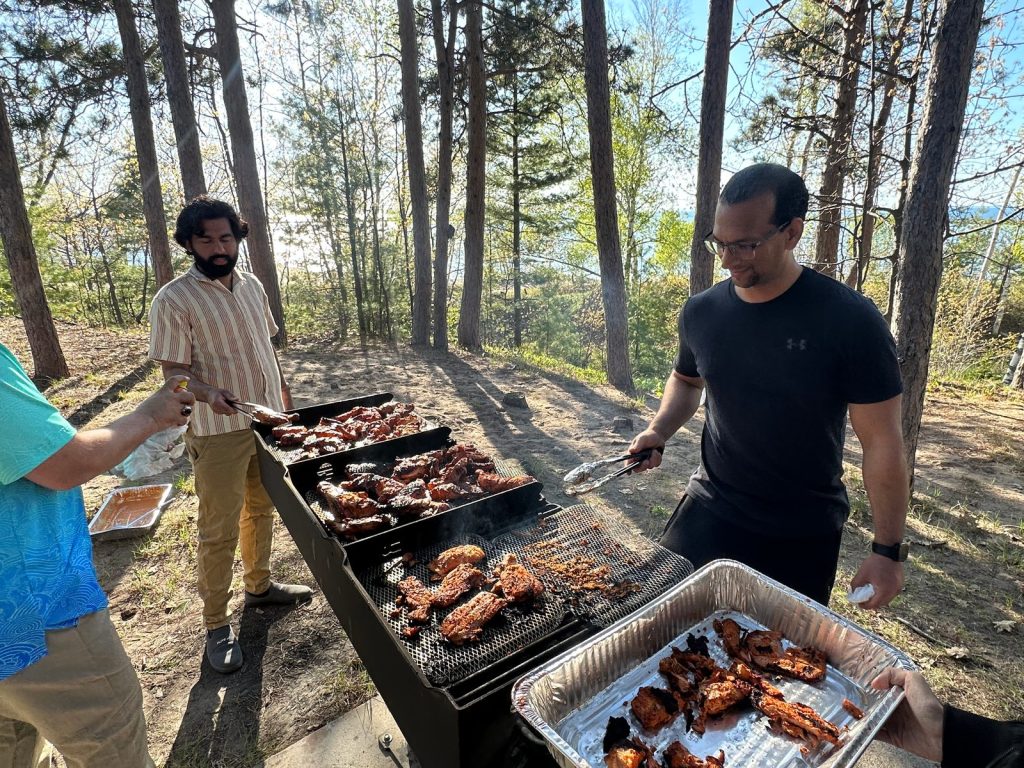Two men turn chicken on a grill while another holds out a foil tray of cooked pieces.