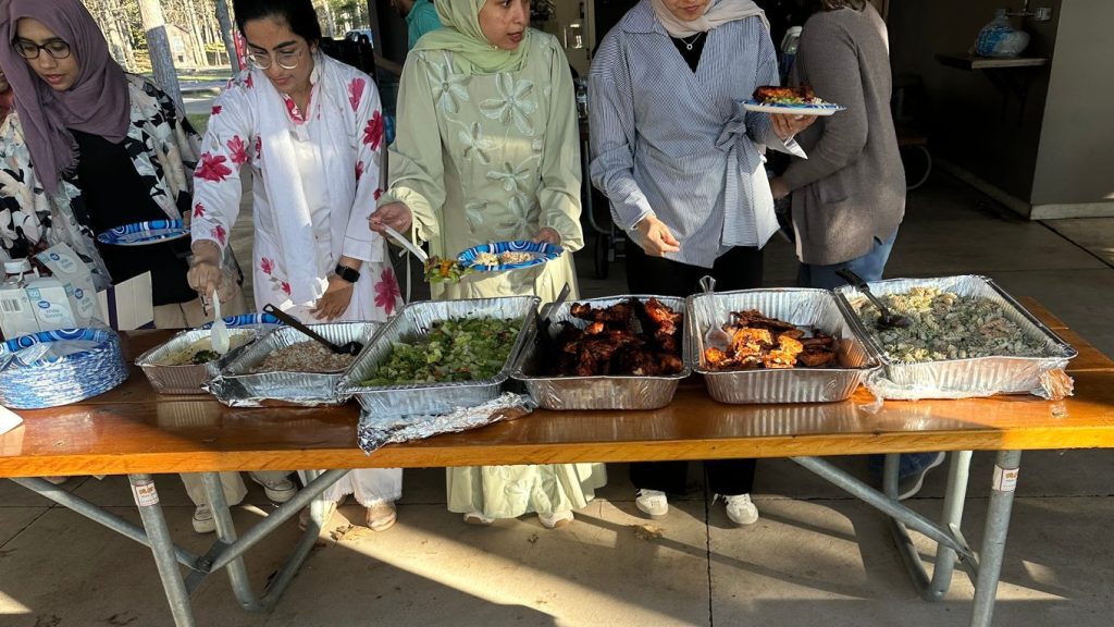 Guests fill their plates from foil trays of rice, salad, grilled chicken and pasta on a picnic table.