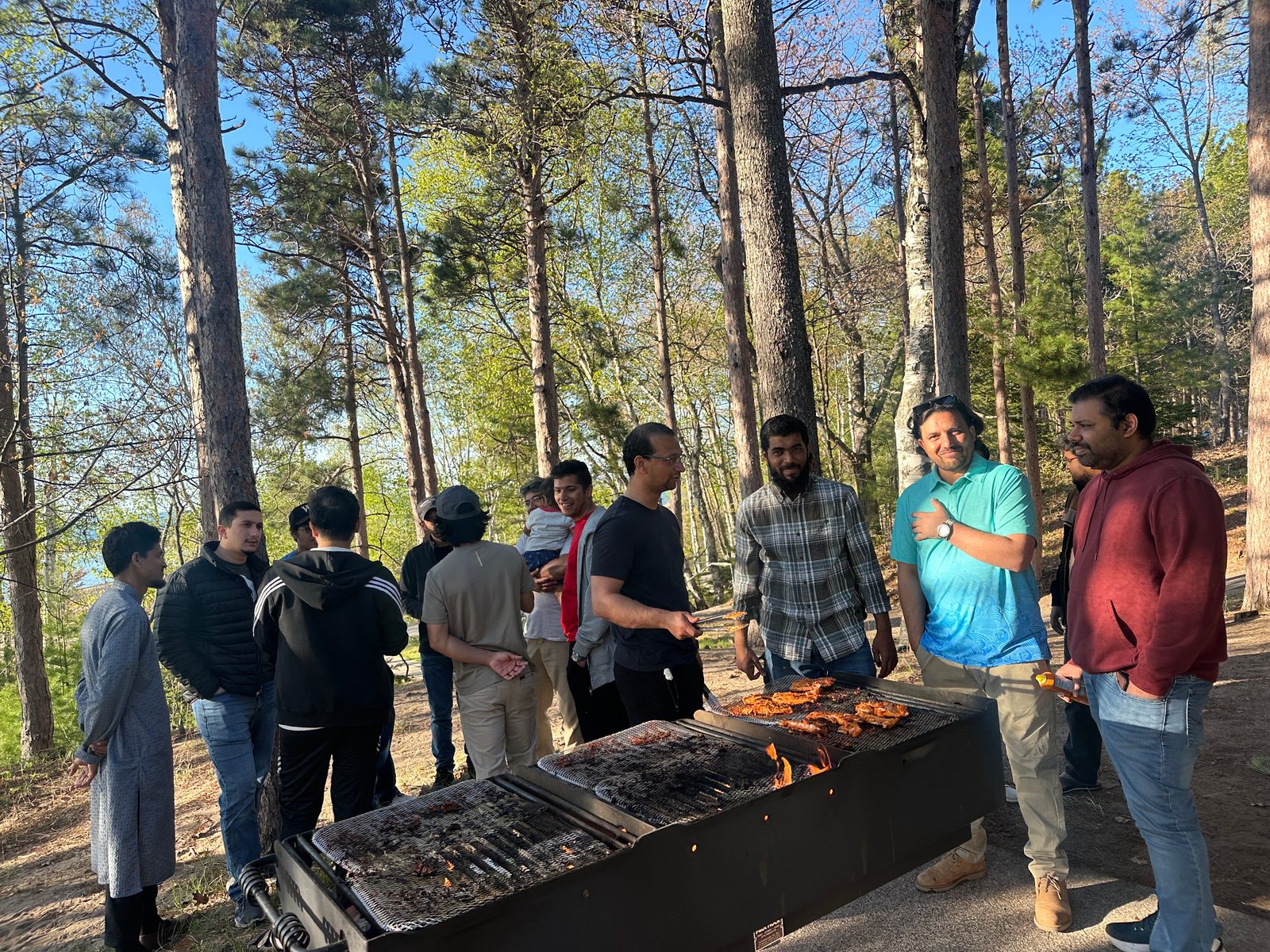 A group gathers around a long charcoal grill of chicken under tall pines.
