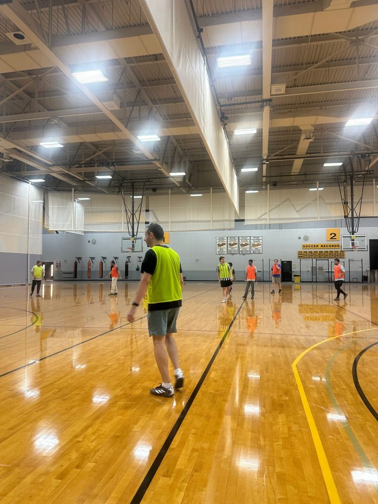 A player in a yellow bib walks across the indoor court as players in orange and yellow bibs spread out.