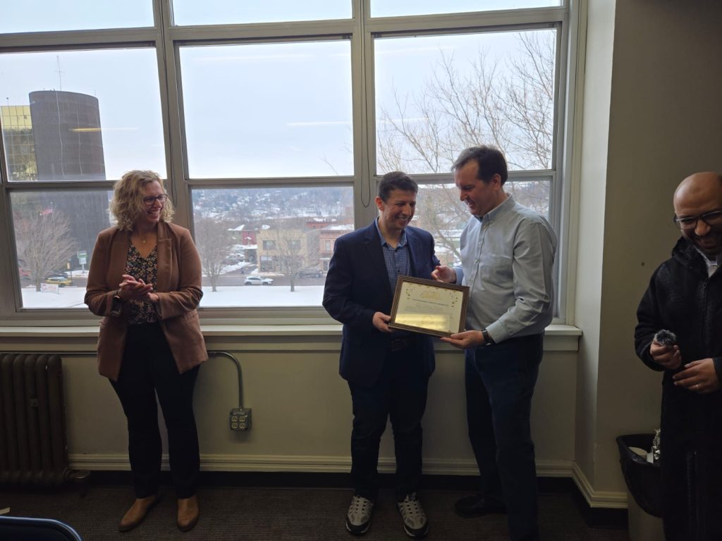 Two men hold a framed award by a window as a woman applauds and another man smiles at the right.