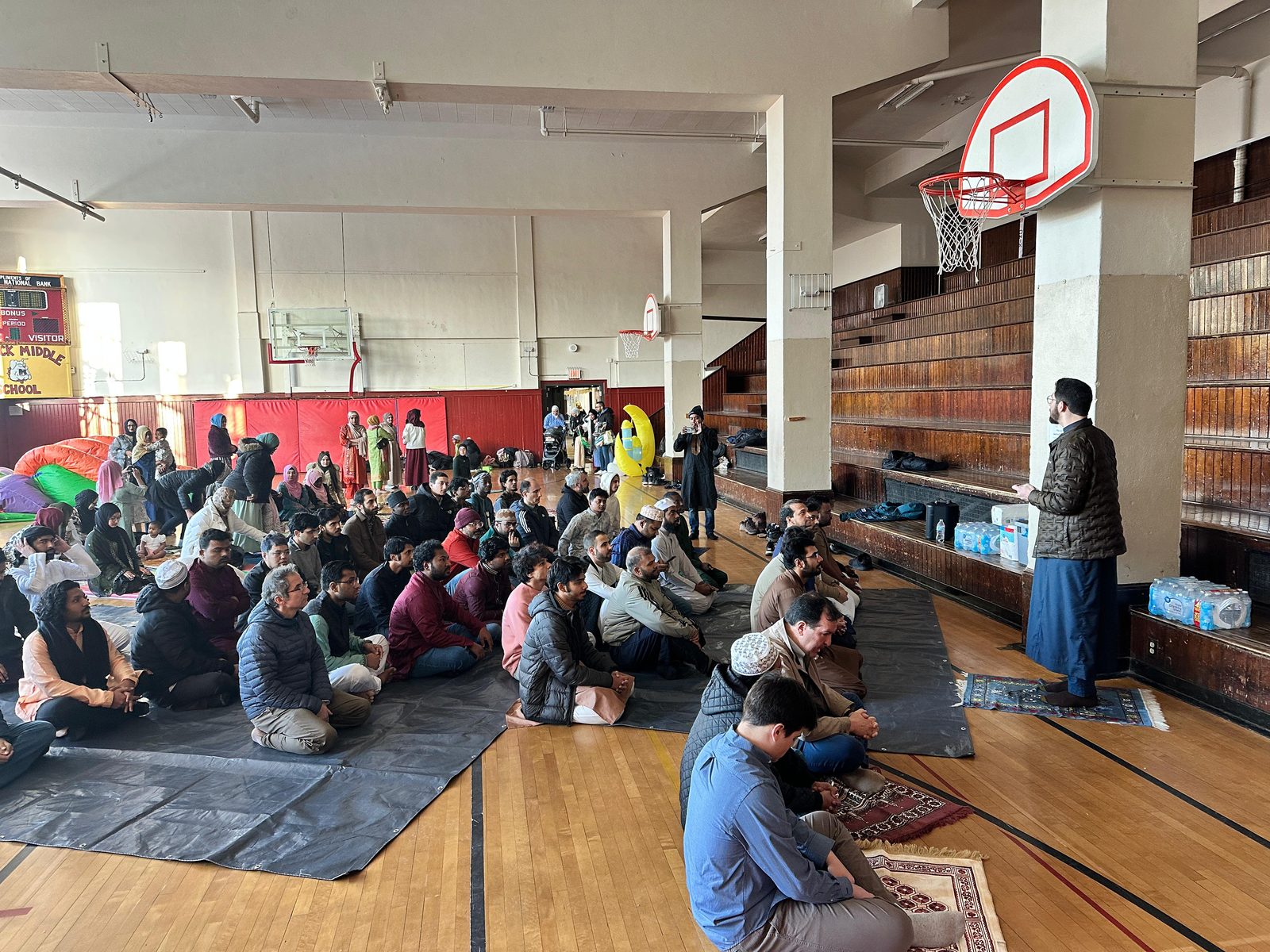 Worshippers sit on tarps in a gym listening to the Eid sermon, with women and children gathered at the back.