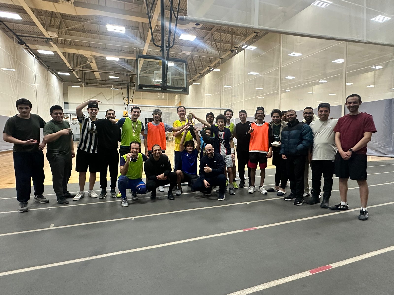 Players in colored bibs and friends pose on an indoor track around a trophy held up in the middle.