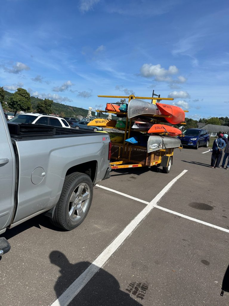 A pickup truck with a trailer stacked with colorful kayaks and canoes is parked in a lot near the waterfront.