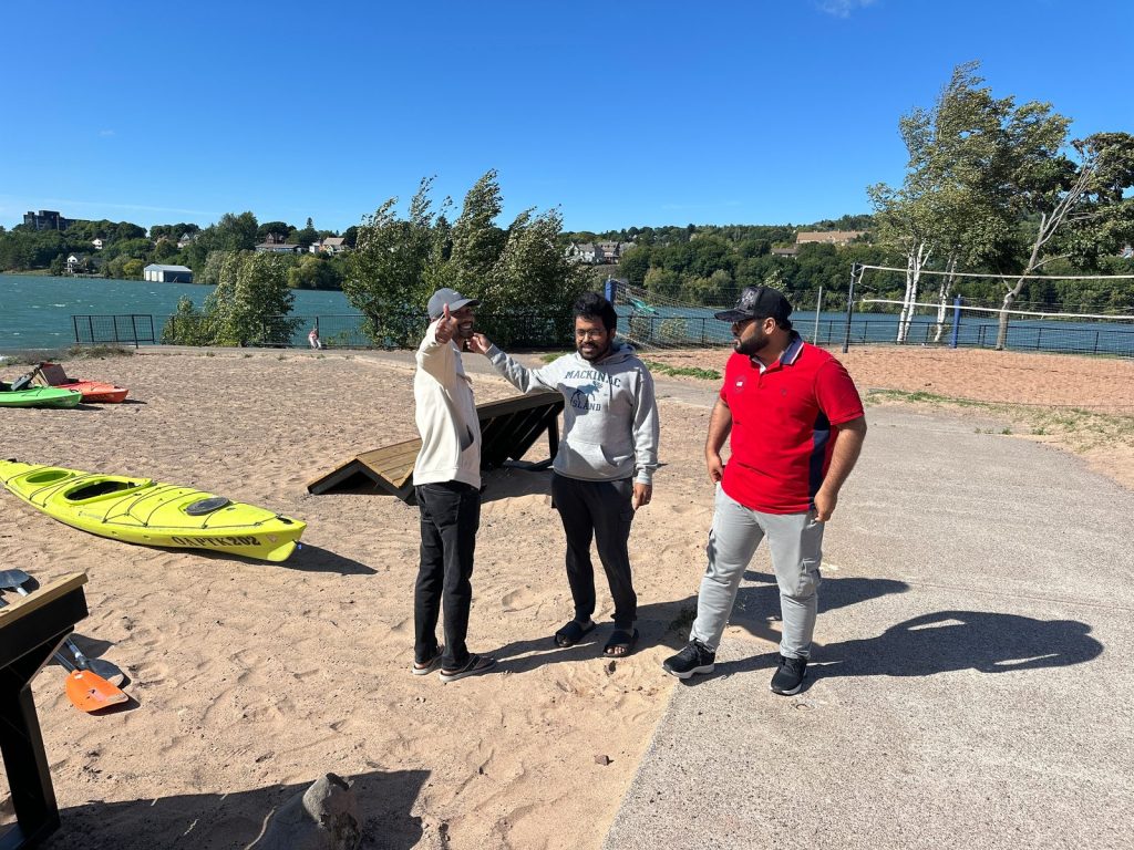 Three men chat on the sandy beach, one giving a thumbs-up, with a kayak beside them and the lake behind.