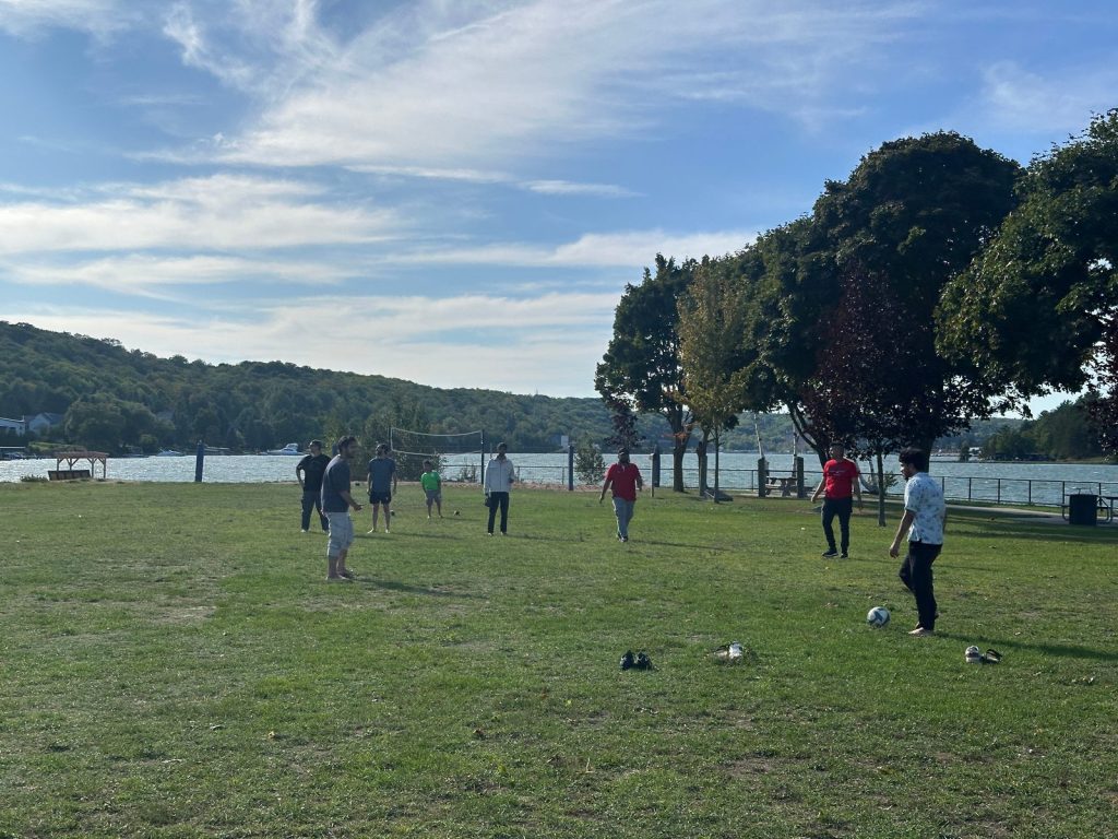A group plays pickup soccer on a grassy lawn beside the lake, with trees and wooded hills behind.
