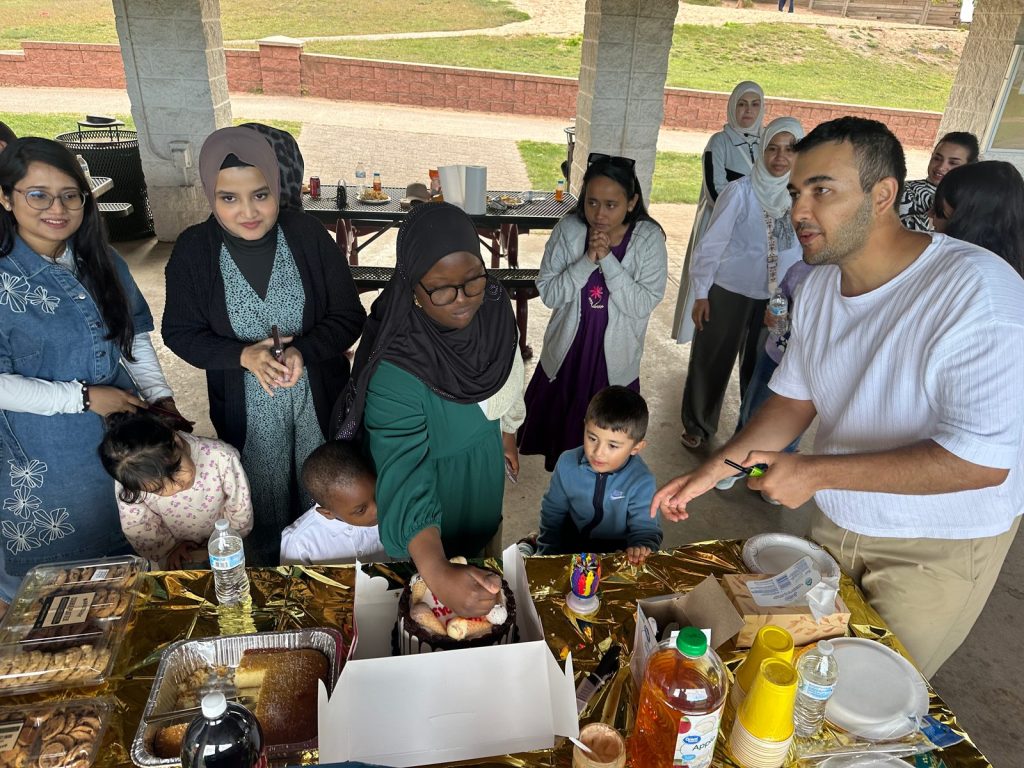 People of all ages gather around a potluck table with cake, cookies and drinks under a pavilion.