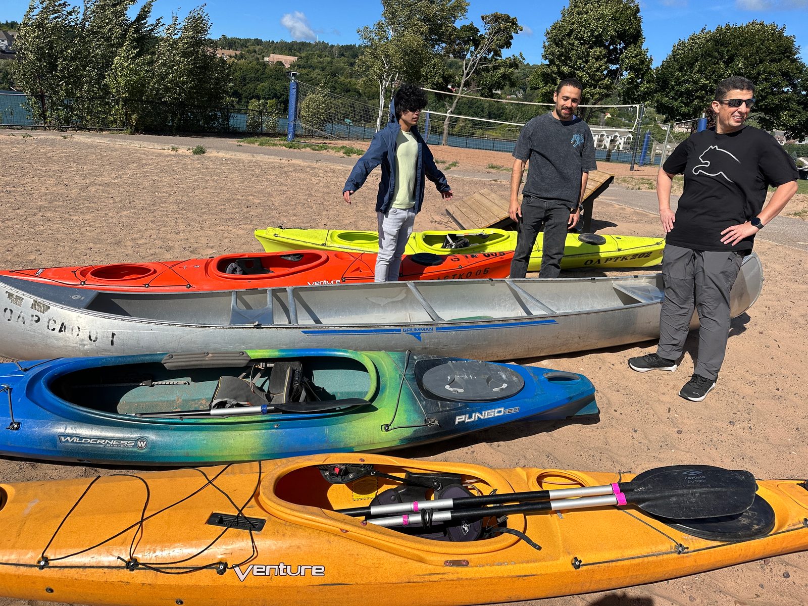 Colorful kayaks and an aluminum canoe lie on the sand as three men stand behind them, with a volleyball net beyond.