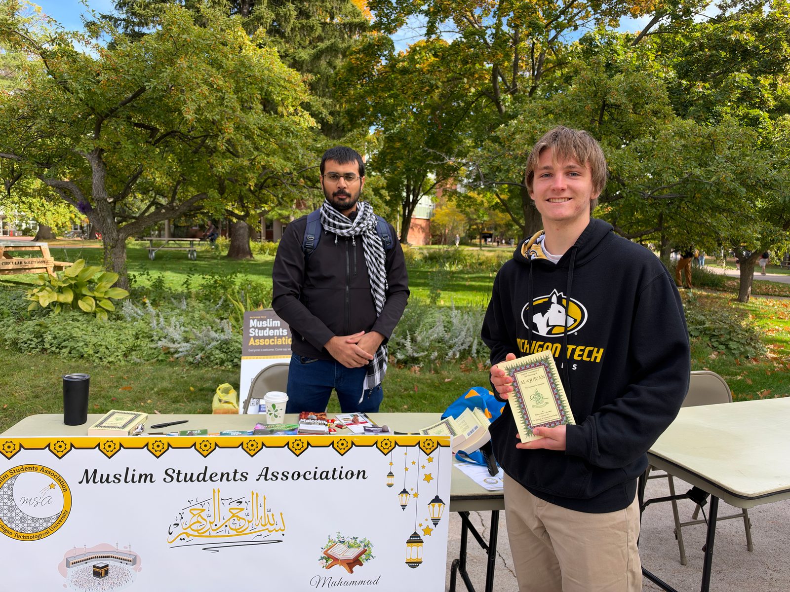 A smiling visitor holds an English translation of the Quran at the MSA table as a man stands behind it.
