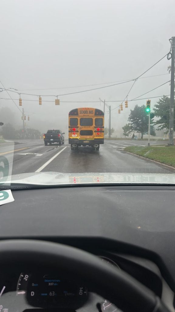 A yellow school bus drives ahead through thick fog at a town intersection, seen through a car windshield.