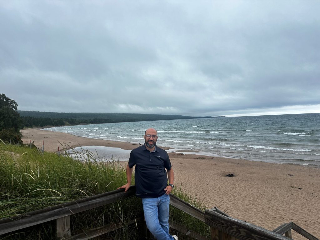 A man leans on a wooden railing above a long, curving sandy beach with waves under an overcast sky.