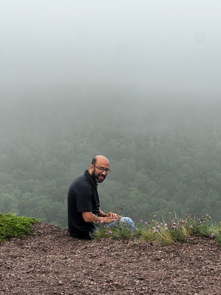 A man sits at the edge of a mountain overlook, with forested slopes below fading into thick fog.