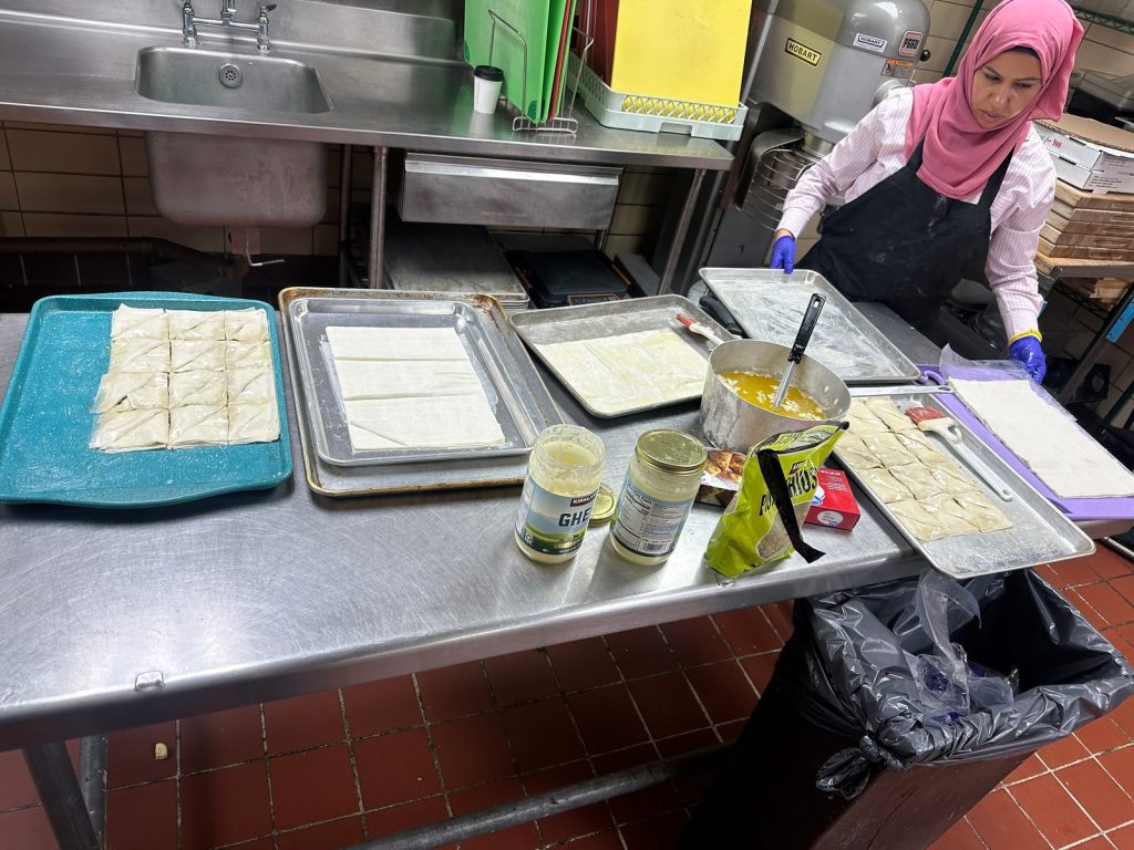 A gloved volunteer in an apron works beside trays of cut phyllo pastry, jars of ghee and pistachios in a kitchen.
