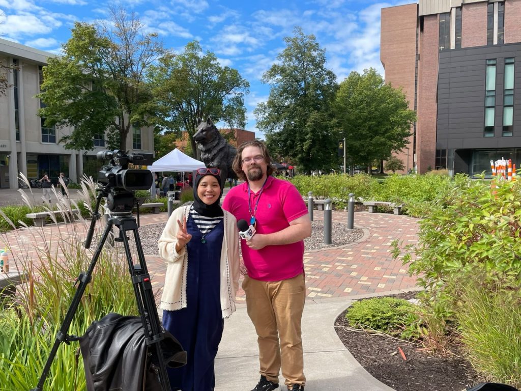A reporter holding a microphone stands beside a smiling woman and a video camera on the Michigan Tech campus.