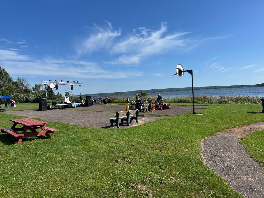 A stage and sound equipment are set up beside the lake before the fair, under a blue sky with wispy clouds.