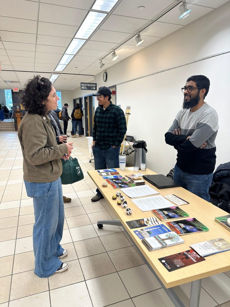 A visitor in an olive jacket chats with two men beside a table of pamphlets, dates and a coffee dispenser.