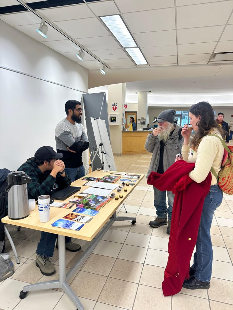 Two visitors sample from small cups in front of the MSA table while two men behind it look on.